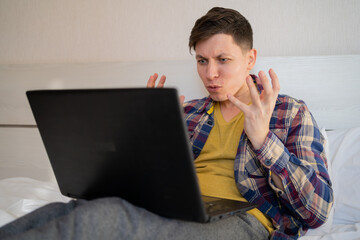 Irritated man sitting on bed looking at laptop screen, arms outstretched, feeling angry and embarrassed, having problems with broken computer, reading bad news in email, lost information,
