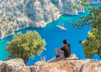 Turkish man sitting at the edge of a cliff and enjoying the beautiful sea scenery. View of Butterfly Valley in Turkey.