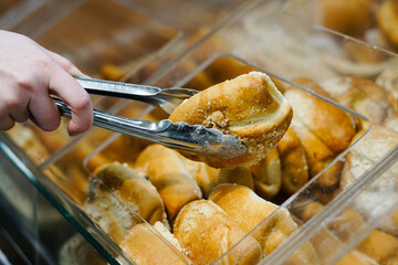 Close up of woman takes fresh baked bun with tongs at a bakery store.