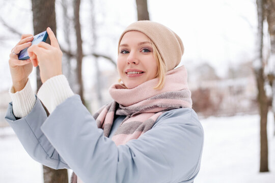 Young blond woman girl in blue warm coat taking photos on smarthone in winter park and smile