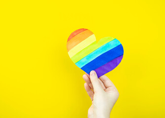 Female hand holding multicolored heart on yellow background. Concept of love , LGBT