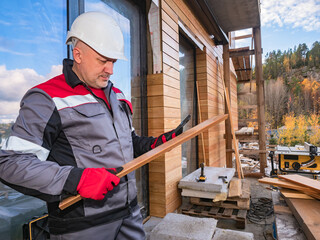 Materials for the construction of the cottage. A man in a gray uniform and construction helmet on a construction site. Foreman orders materials for the exterior decoration of the cottage by phone.
