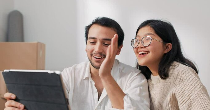 Lovers are sitting on floor in newly bought apartment elegantly dressed man is holding tablet couple is chatting with parents friends over internet waving to camera bragging about moving into flat