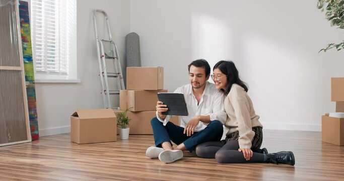 Lovers are sitting on floor in newly bought apartment elegantly dressed man is holding tablet couple is chatting with parents friends over internet waving to camera bragging about moving into flat