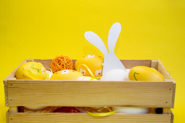 Colorful Easter eggs in a wicker basket and a wooden box on a yellow, green background. Space for...