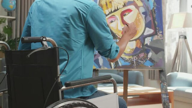 Hind View Of An Asian Artist Man In Wheelchair Holding Paintbrush And Mixing It With Colour Before Painting A Girl'S Face On The Canvas
