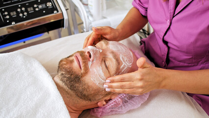 Obraz na płótnie Canvas Cosmetologist makes a man a facial therapeutic. Middle-aged man in a spa for beauty treatments. Beautician extrude from bottle with moisturizing cream on his hand to apply it to the male face.