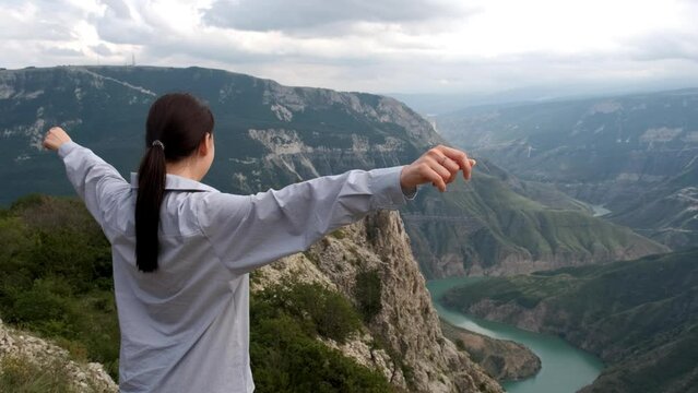 A caucasian brunette girl in a shirt stands on the edge of the mountain above the great Sulak Canyon and raises her hands up. Tourism, travel