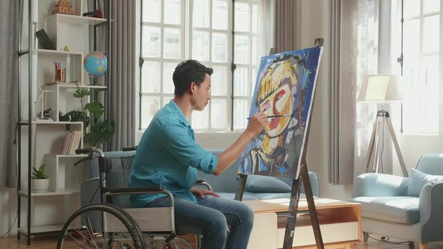 Hind View Side View Of An Asian Artist Man In Wheelchair Holding Paintbrush Mixed Colour And Painting A Girl On The Canvas
