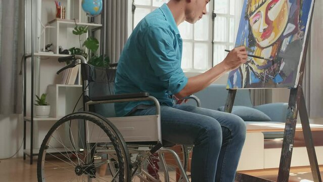 Medium Shot Side View Of An Asian Artist Man In Wheelchair Holding Paintbrush Mixed Colour And Painting A Girl On The Canvas
