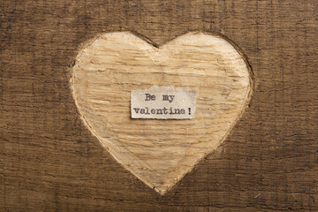 Be my Valentine - tiny typed text note close up. Valentines Day greetings concept. Carved heart shape on wood as background for Valentines greeting card.
