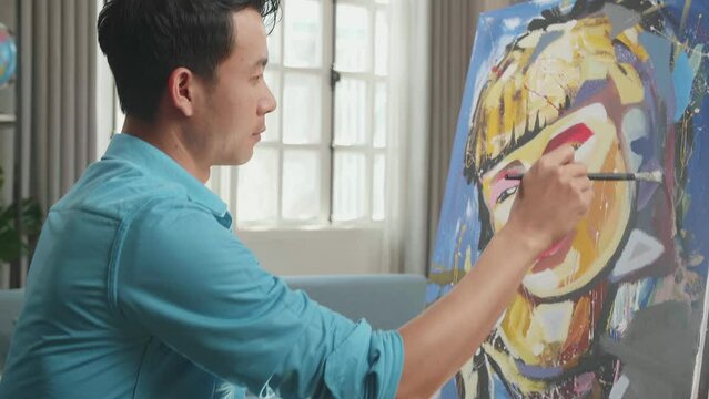 Side View Of An Asian Artist Man In Wheelchair Holding Paintbrush Mixed Colour Turn To Smile While Painting A Girl On The Canvas
