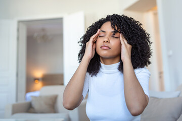Fototapeta na wymiar Portrait of a young black girl sitting on the couch at home with a headache and pain. Beautiful woman suffering from chronic daily headaches. Sad woman holding her head because sinus pain