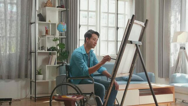 Side View Of An Asian Artist Man In Wheelchair Holding Paintbrush Mixed Colour And Thinking Before Painting On The Canvas
