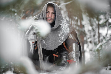 A knight in the armor with the sword and red gerbera flower in the winter snowy forest. Medieval...