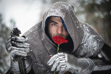 A knight in the armor with the sword and red gerbera flower in the winter snowy forest. Medieval...
