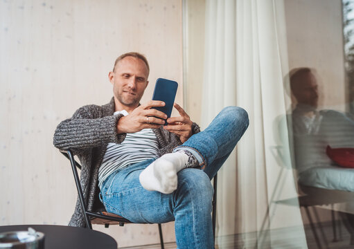 Smiling middle-aged man dressed open cardigan, jeans, and warm socks sitting on house balcony and smiling when he using modern smartphone. Everyday lifestyle photo with modern devices concept image..