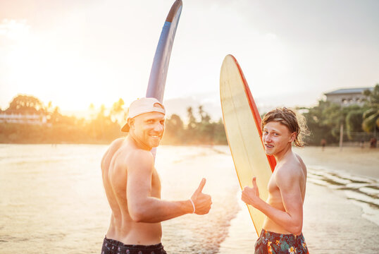 Father with teenager son standing with surfboards on the sandy ocean beach with palm trees on background lightened with sunset sun. They smiling and showing thumbs up. Family active vacation concept