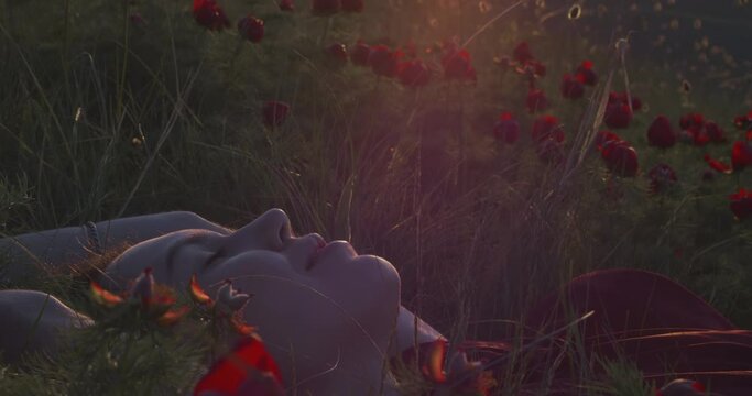 Slow motion. Portrait of a sexy woman in profile who lies in the grass against a background of wild peonies in the rays of the sunset