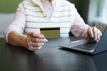 Hand of contemporary elderly woman with credit card sitting by table in front of laptop and...