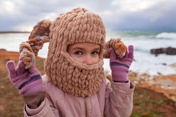 A girl in a fashionable knitted balaclava walks on the shore of the winter sea. Child wearing a...