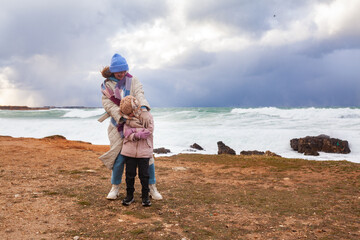Happy mom and daughter are walking on the shore of a winter stormy sea, a woman and a girl are...