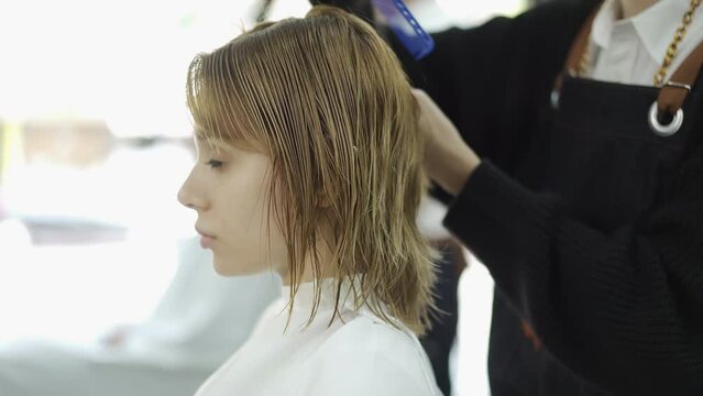 Caucasian female client in haircut cape sitting on hairdressing chair looking in the mirror while having a haircut by Asian female hairdresser in salon.