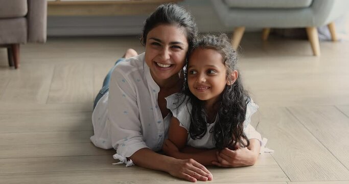 Beautiful mother and little daughter portrait, homeowner family, tenancy concept. Indian woman hugging cute kid lying together on warm floor with underfloor heat system smile look at camera feel happy