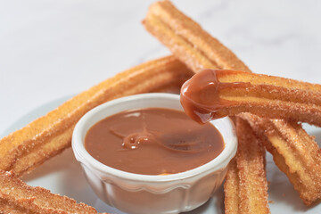 Traditional churros with sugar and cinnamon served on a marble table, with a bowl of caramel...