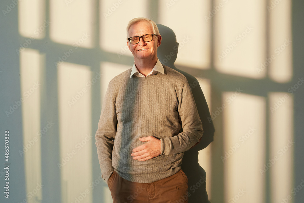 Wall mural smiling elderly man in brown pants and pullover and eyeglasses standing against sunlit wall with sha