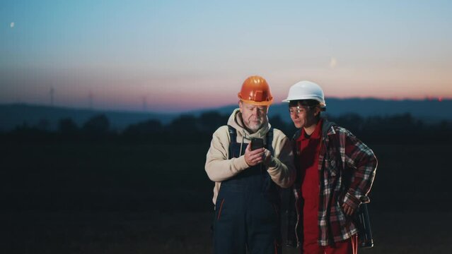 Smiling two engineers woman and mature man in hard hat use phone to check inspecting wind turbine power station in the evening. Slow motion