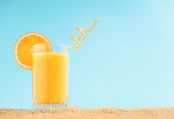 Selective focus of glass with orange fruit organic juice served with citrus slice and straw full of vitamins on sandy beach against sky blue background. Summer drinks concept. Image with copy space