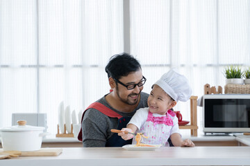 Asian young father with beard and little cute mixed race daughter with apron and chef hat cooking and enjoy eating spaghetti together in kitchen at home. Family activity in special day concept