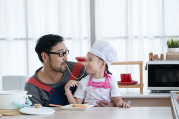 Asian young father with beard and little cute daughter with apron and chef hat cooking together and enjoy eating spaghetti same string in kitchen at home. Family activity in special day concept