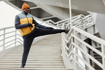 Young contemporary sportsman of African ethnicity standing on staircase during physical exercise with one leg stretched