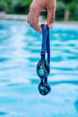 Man`s hand holding professional swimming goggles with pool blue water on background