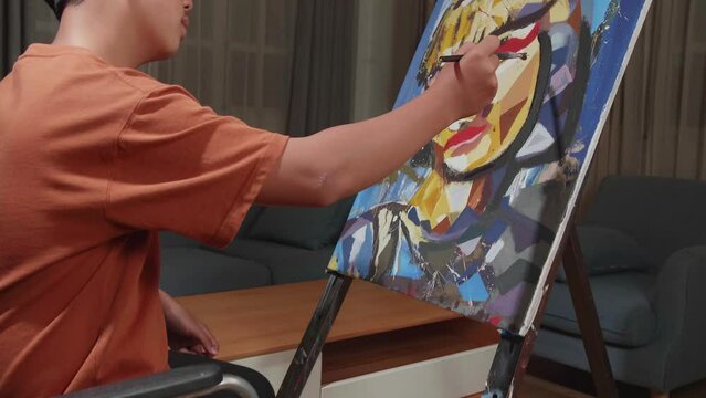 Asian Artist Boy In Wheelchair Holding Paintbrush And Mixing It With Colour Before Painting A Girl'S Face On The Canvas

