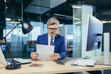Experienced gray-haired businessman working at a computer, paperwork in the office, reading a letter from the bank