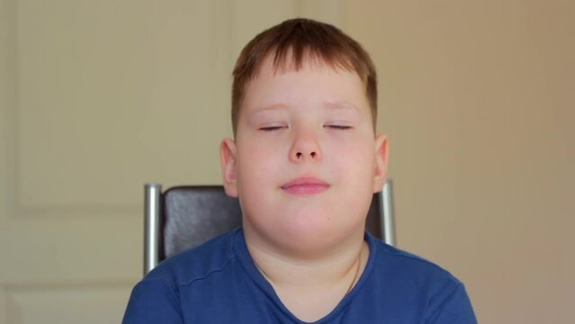 A Fat Boy Sits On A Chair And Yawns. He's Wearing A Blue T-shirt.