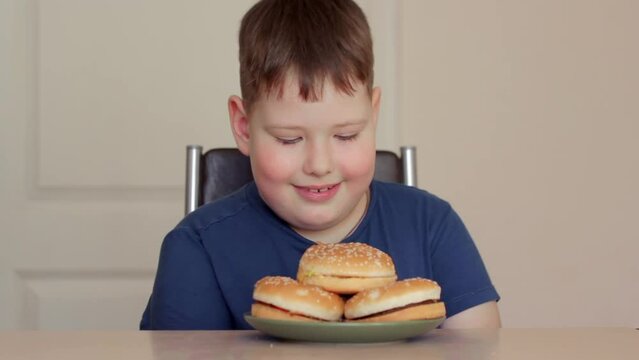A fat boy is sitting on a chair at the table and looking at hamburgers. He smiles and wants to eat.