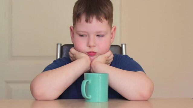 A fat child is sitting at a table with his chin in his hands, looking away and smiling. There is a green mug on the table.