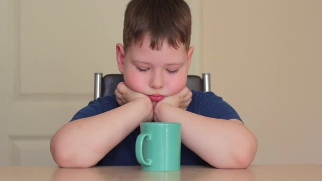 There is a green mug on the table. A fat child is sitting at the table, propping his chin on his hands, looking at a mug.