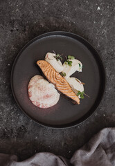 Piece of grilled salmon with sauce, mashed potatoes and microgreens on a black round plate