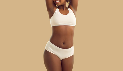 Happy young black woman with curvy body posing in underwear. Beautiful plus size female model wearing basic bra and underpants standing hands behind head on solid beige color background, cropped shot