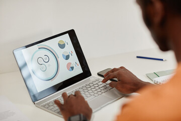 African American male economist or accountant sitting in front of laptop with financial data on screen and analyzing diagrams