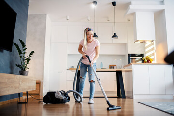 A smiling woman vacuuming living room floor at her home. - obrazy, fototapety, plakaty