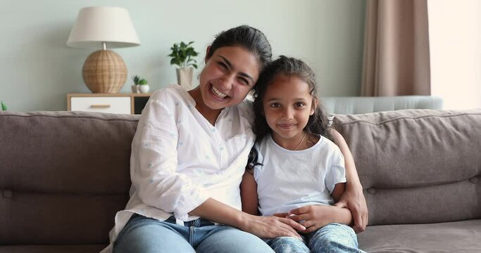 Cheery woman hugs her little 6s daughter sit together on sofa at home laughing look at camera enjoy video conference call. Indian family bond, affection, happy motherhood, life value, cherish concept