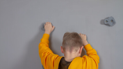 A boy in a yellow hoodie climbs a gray wall on rock climbing stones, a child learns rock climbing...
