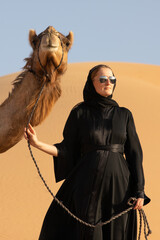 Young woman in Emirati national dress (abaya) with a camel in Empty Quarters desert dunes. Abu Dhabi, UAE.