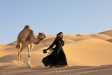 Young woman in Emirati national dress (abaya) with a camel in Empty Quarters desert dunes. Abu Dhabi, UAE.